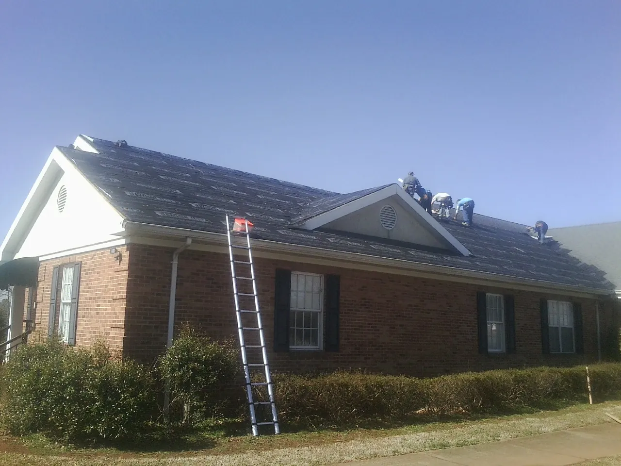 Crew tearing shingles off a brick ranch roof with a ladder set — Emergency Roof Repair work by our crew in Ira